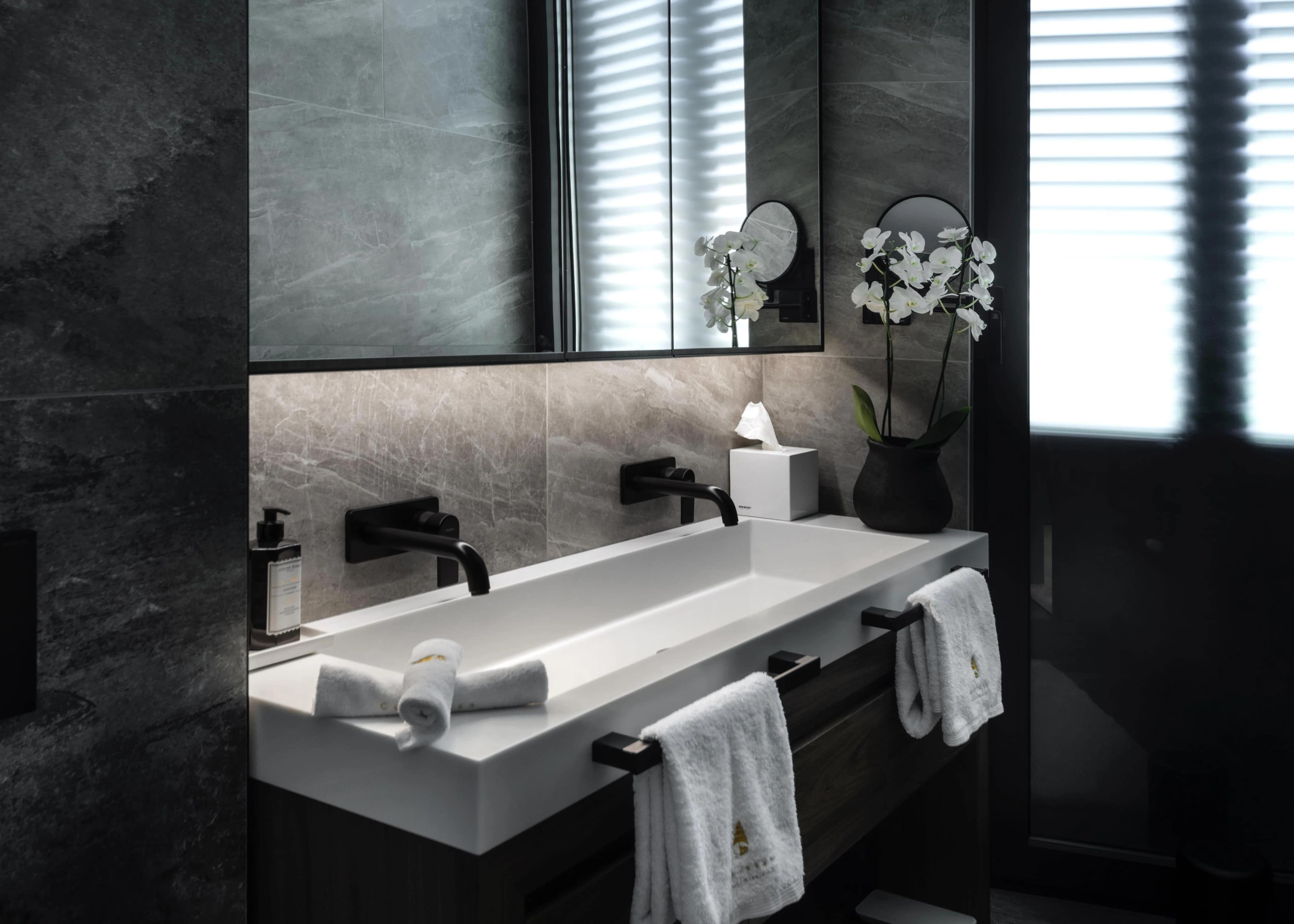 Spiridon bathroom — the trough basin in dark stone, black fixtures, an orchid