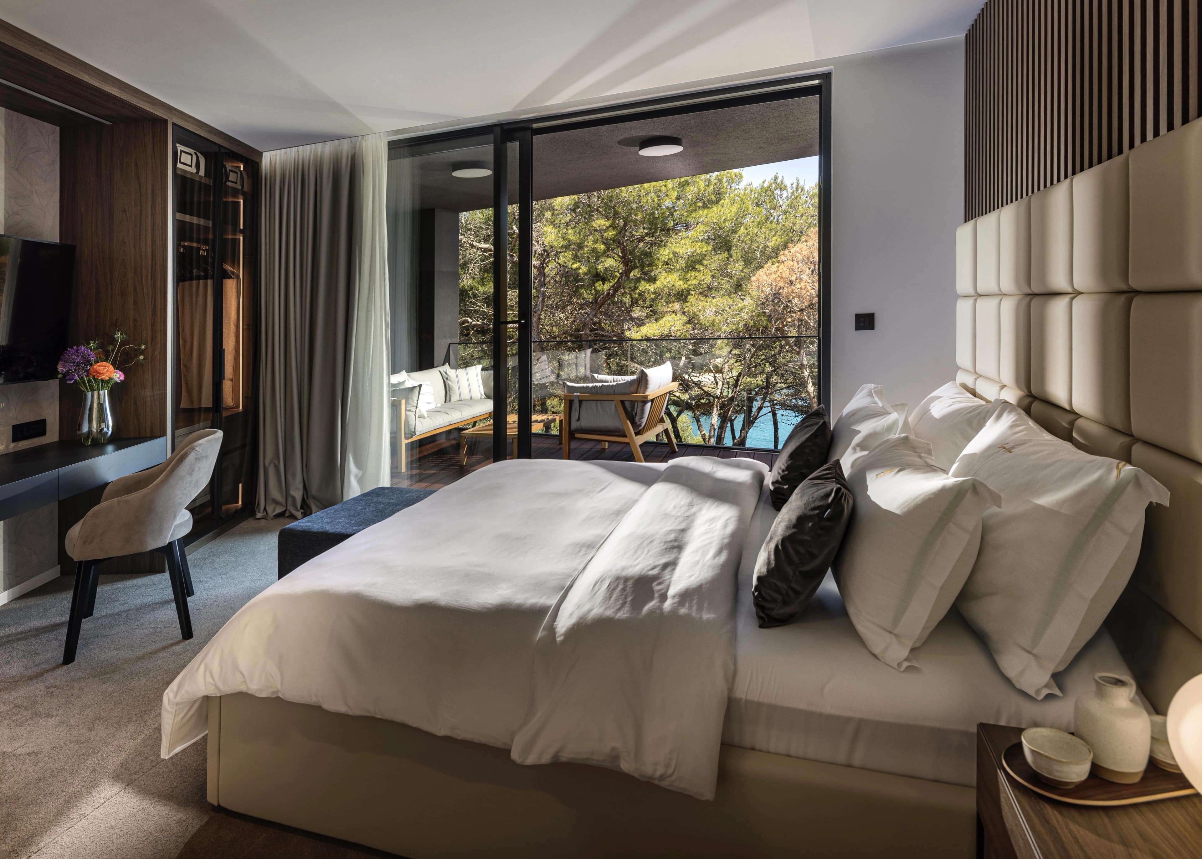 Spiridon suite — beige padded headboard, built-in TV wall, vanity desk and a balcony lounge through the sliding doors