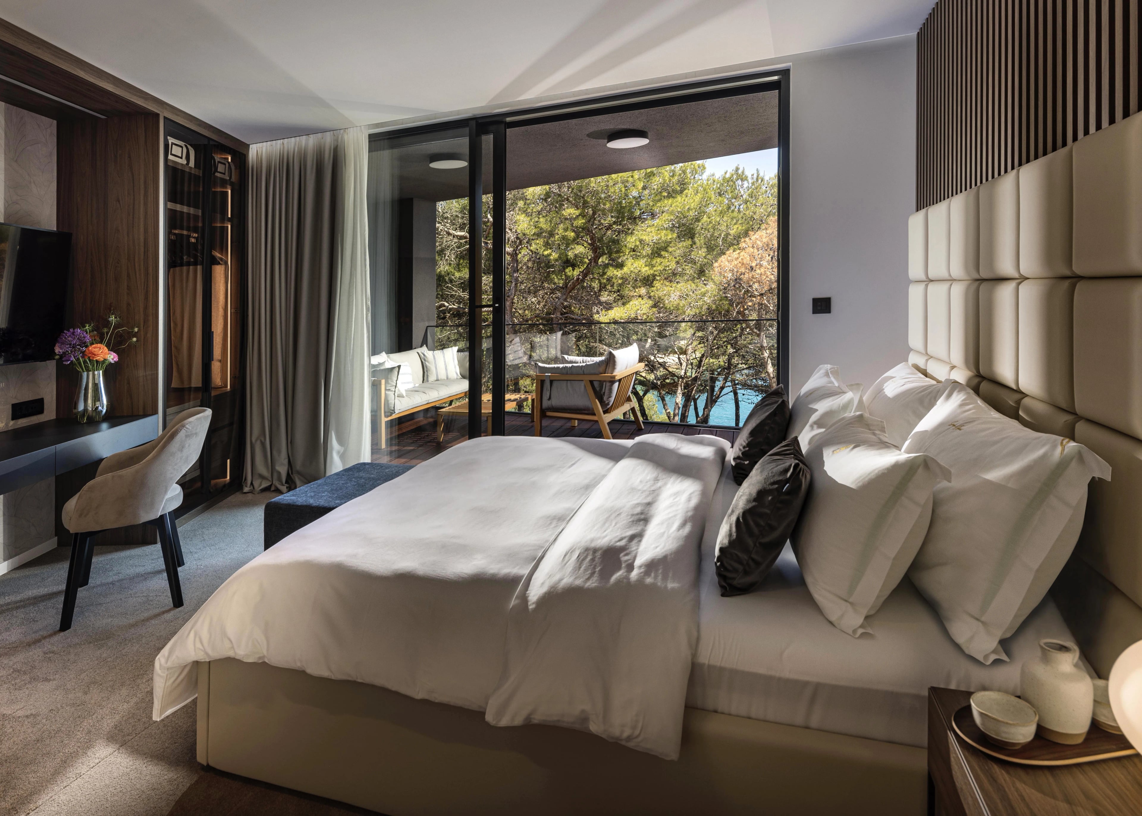 Spiridon suite — beige padded headboard, built-in TV wall, vanity desk and a balcony lounge through the sliding doors