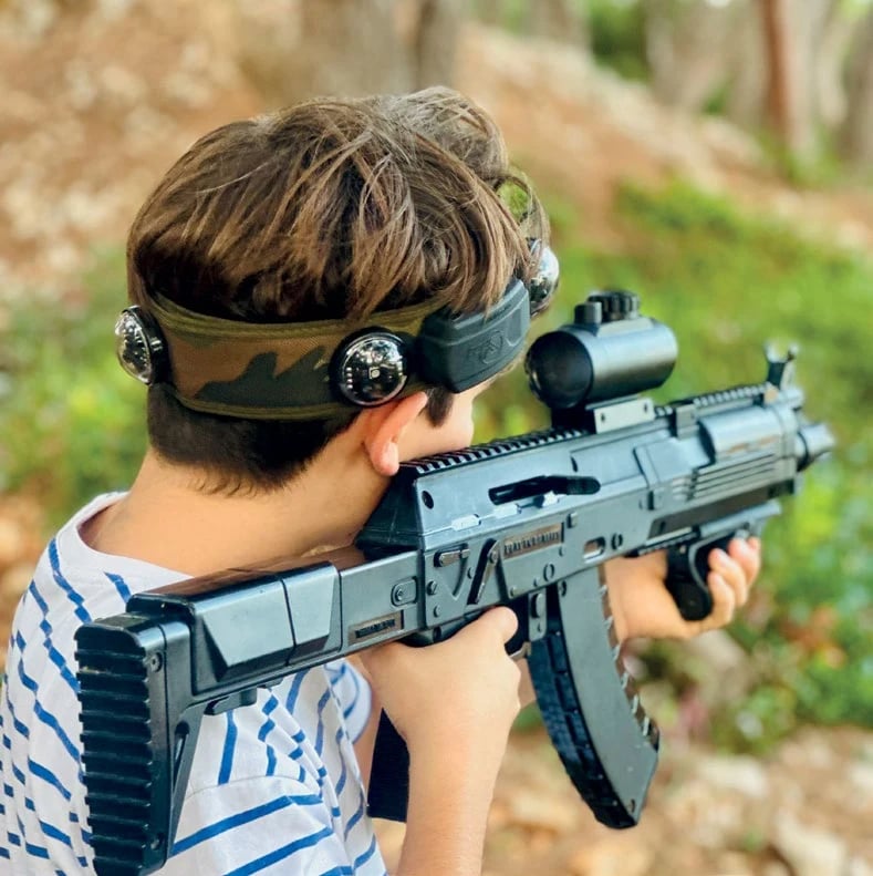 A boy in a camo headband aiming a laser-tag rifle in the pine woods