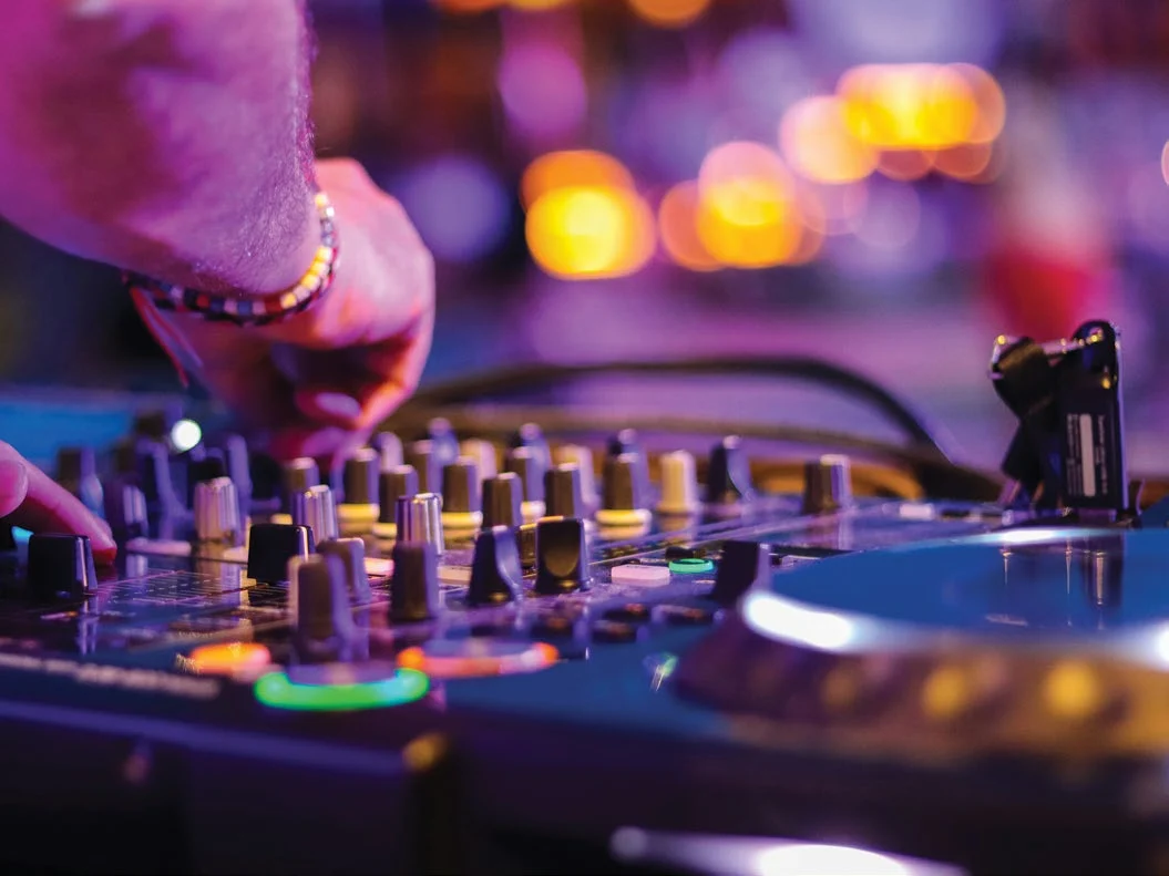 A DJ’s hands on a mixer with coloured lights blurred behind it