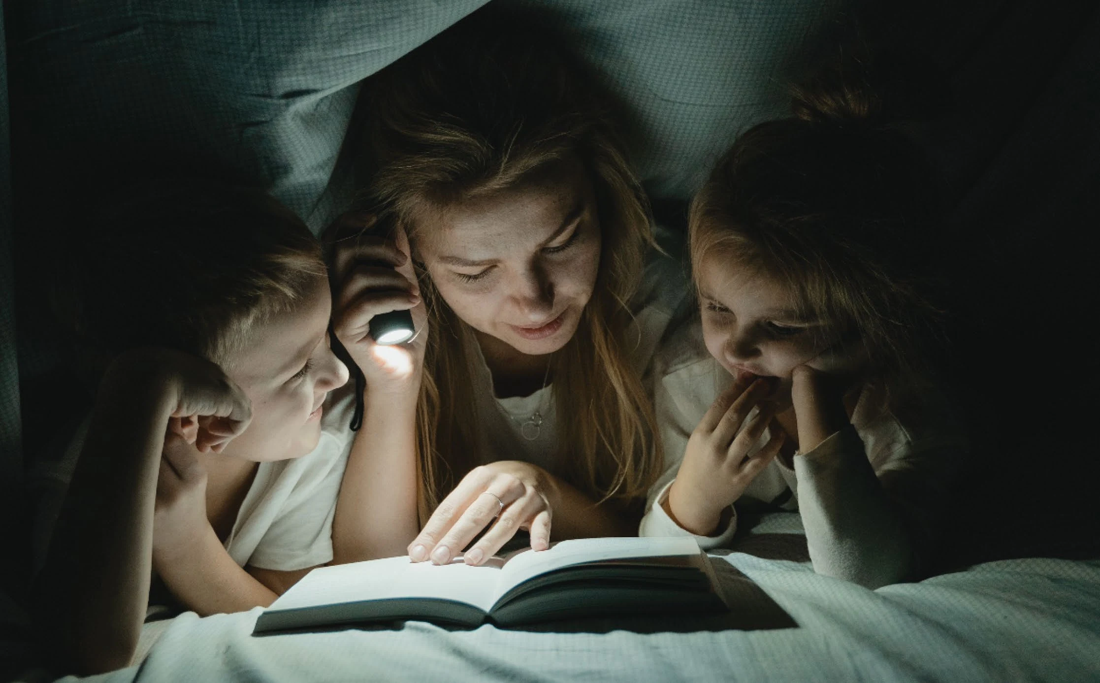 Two children and their carer reading together under a blanket by flashlight
