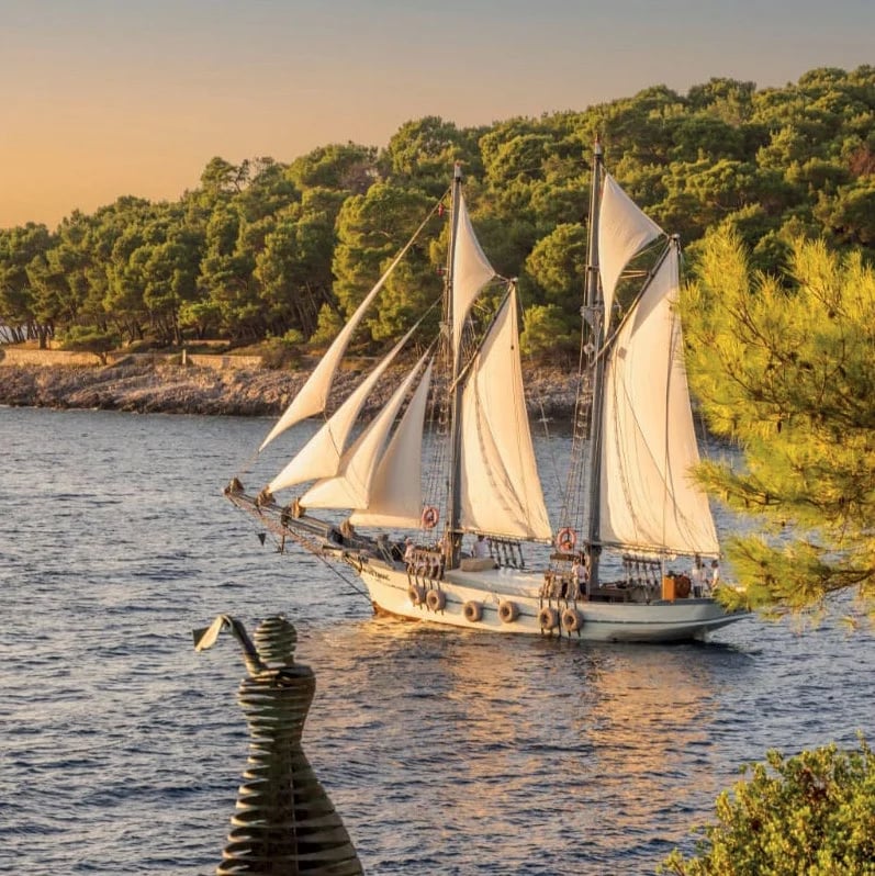 The Nerezinac historic sailing vessel passing a pine-covered shoreline at golden hour