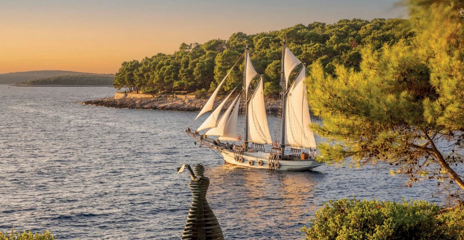 A two-masted sailing schooner gliding past a pine-covered shore at golden hour