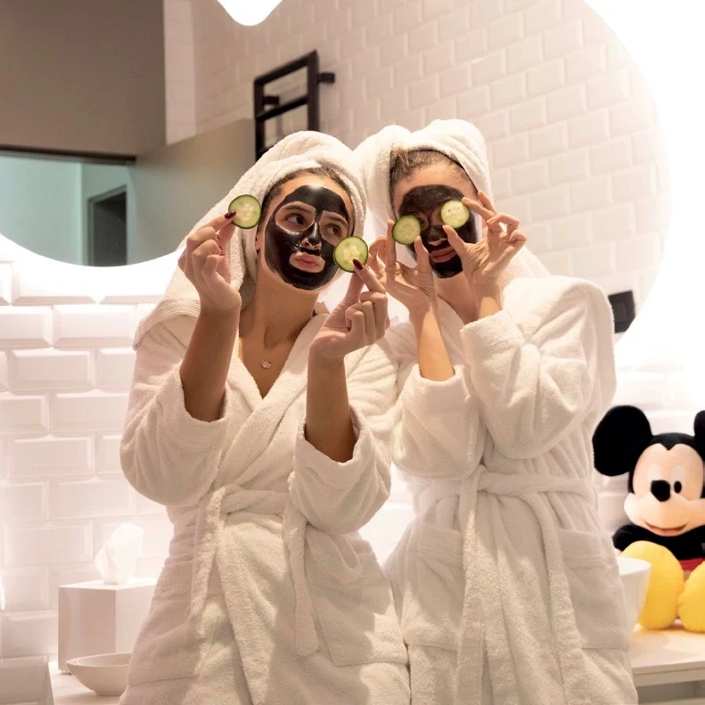 Two girls in white robes with cucumber eye-pads and a Mickey Mouse doll