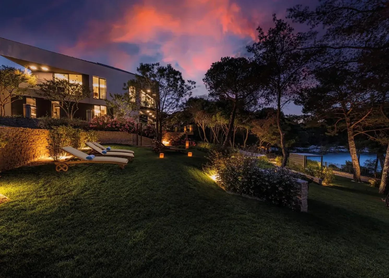 Loungers on the lawn under a pink dusk sky, the villa lit behind